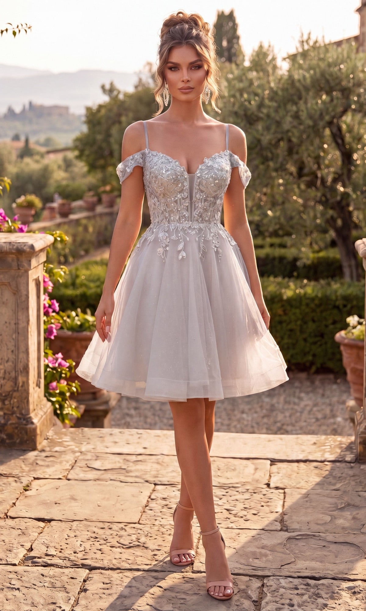 A woman in the Short Homecoming Dress: Chicas C359, featuring a silver off-shoulder design with floral lace appliques and a tulle skirt, stands outdoors on a stone path surrounded by greenery.
